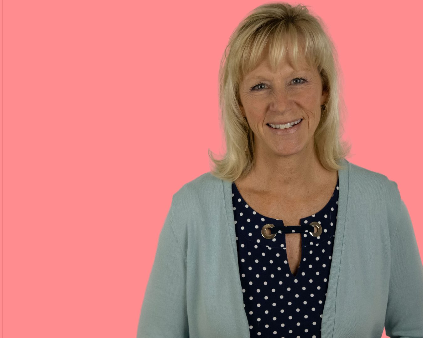 Patti Kinnear smiling in a polka dot shirt and blue cardigan. The background appears pink